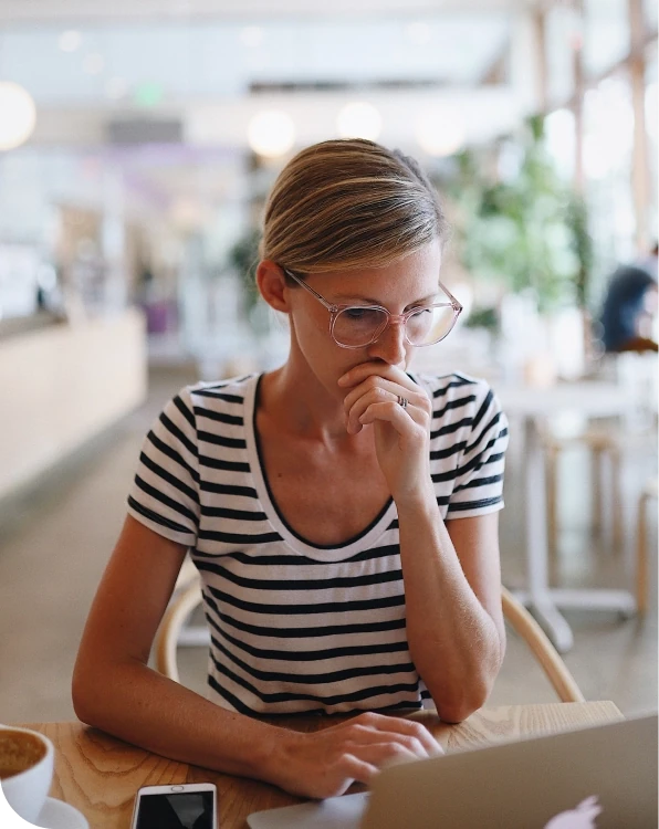 Une personne assise à une table de café, regardant pensivement un écran d'ordinateur portable avec une main près de la bouche, suggérant une préoccupation ou une prise de décision en travaillant en ligne.