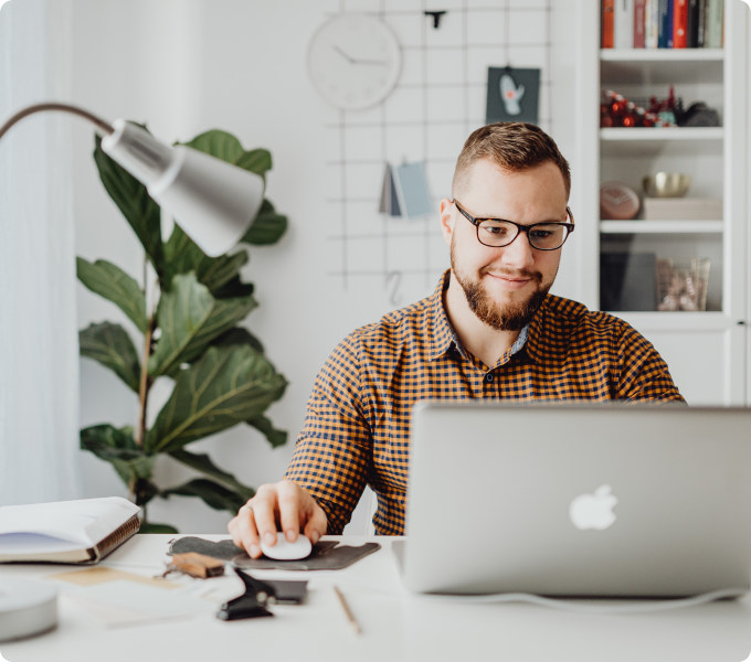 Person working at a laptop in a modern home office with a desk lamp, plant, and bookshelf in the background.